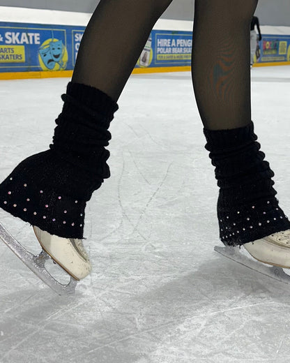 A pair of legs wearing black leg warmers with a sparkly design, on an ice skating rink.